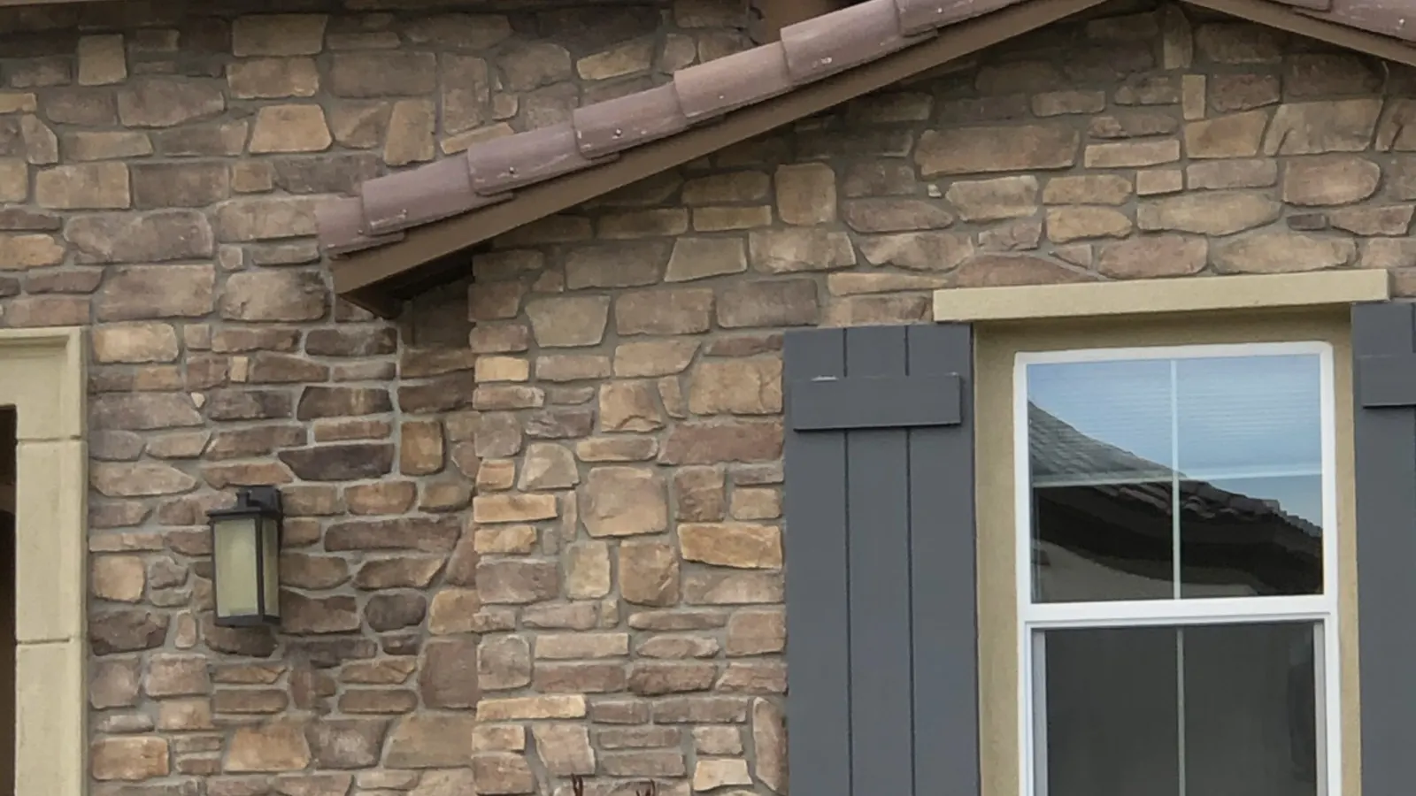 Manufactured stone veneer covers a single-story home's exterior around a shuttered window, beneath a clay tile roof.